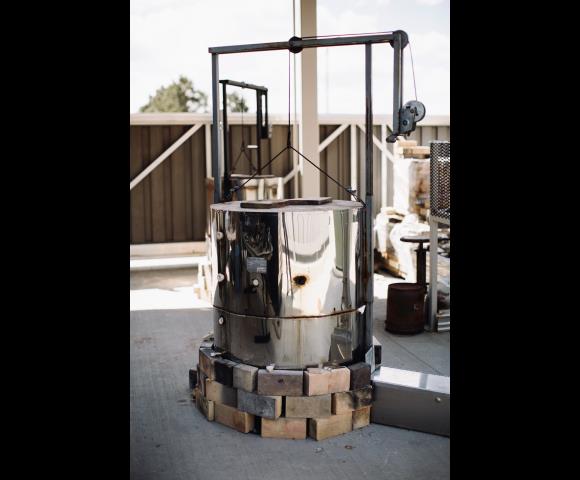 Outdoor top-loading electric kiln on a base of fire bricks with a metal lifting frame above it.
